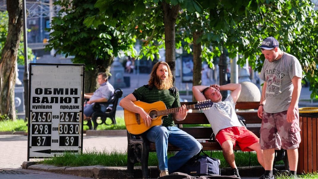 Вуличний музикант грає на гітарі, сидячи на лавці біля стенду з курсом валют, в Києві, 11 червня 2020 року