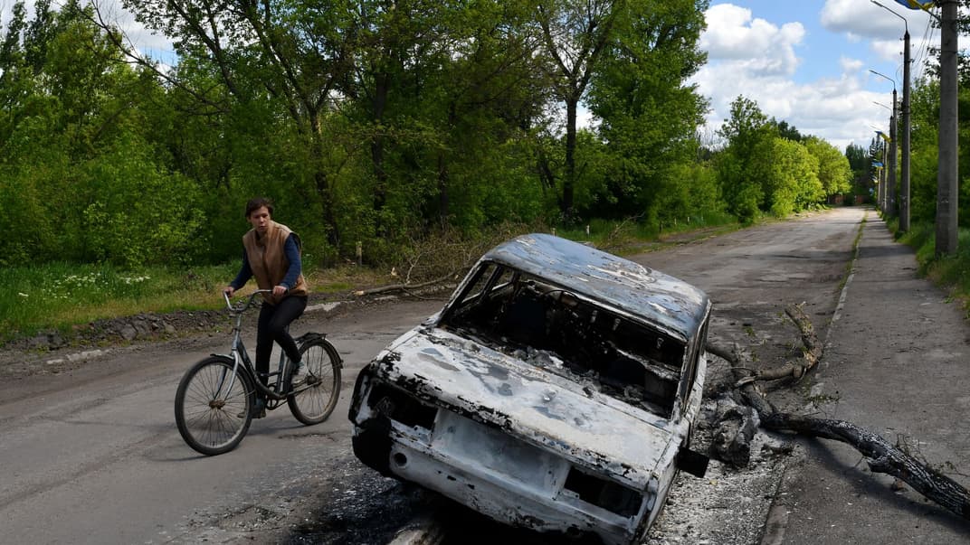 Чоловік проїжджає на велосипеді повз знищений обстрілом автомобіль на вулиці в селі Ню-Йорк Донецької області, 16 травня 2022 року