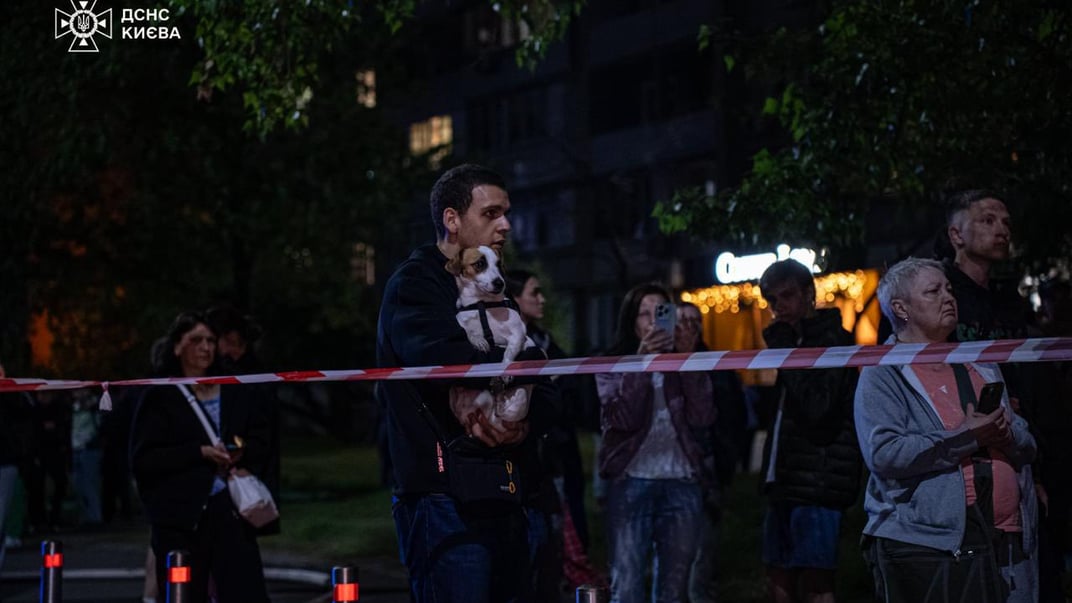 Люди стоять на вулиці під багатоповерхівкою, яка зайнялася після російської дронової атаки по Києву, 4 травня 2025 року