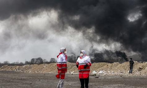 Представниці іранського Товариства Червоного Півмісяця стоять на тлі диму після атаки по нафтопереробному заводу «Шахран», 8 березня 2026 року