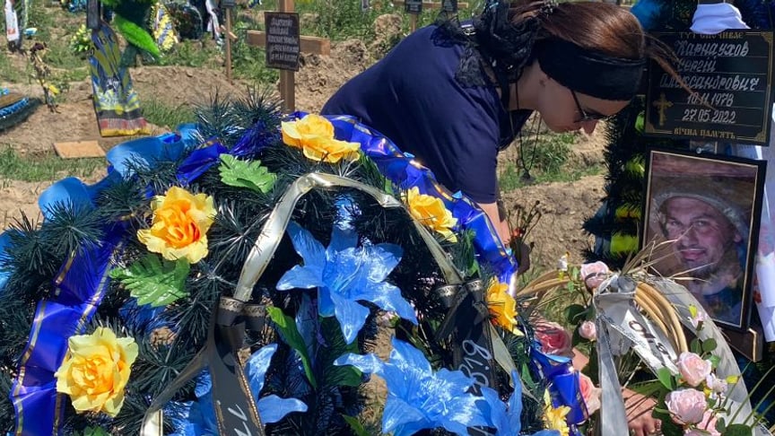 Алина ставит в вазочку живые цветы между пластиковыми венками на могиле своего мужа